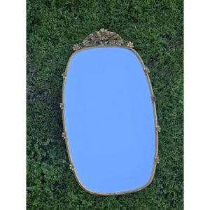 May include: An oval-shaped gold-framed mirror with floral accents. The mirror has a light blue reflection and is surrounded by a decorative gold frame with small flower details. The top of the frame features a larger floral design.