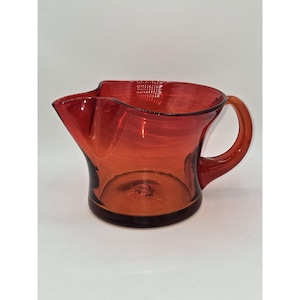 May include: A translucent red glass pitcher featuring a wide, flared rim with a prominent pouring spout and a curved handle on the side. The vessel has a smooth, rounded body with a circular base, showcasing a deep crimson hue throughout.