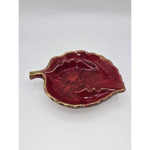 May include: A decorative leaf-shaped dish with a deep red interior and a gold-colored rim. The dish has a textured surface and a glossy finish, adding a touch of elegance. This unique piece is perfect for serving small items or as a decorative accent.