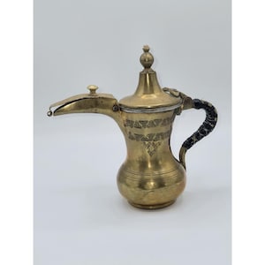 May include: A vintage brass coffee pot with a long spout and a decorative handle. The pot features etched patterns around the body and a small lid. The handle is wrapped in a dark material. The pot is a warm gold colour.
