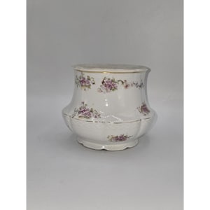 Könnte beinhalten: Ein weißer Keramikbehälter mit Deckel, verziert mit einem Blumenmuster in Lila- und Grüntönen. Der Rand und die Kanten sind mit goldenen Zierleisten versehen. Der Behälter hat eine abgerundete Form.