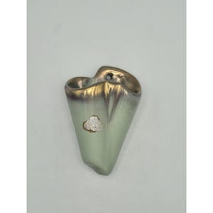 Puede incluir: Un jarrón de bolsillo de pared vintage con una forma única y una combinación de colores verde claro y dorado. El jarrón tiene una pequeña etiqueta con texto. El borde superior es dorado y el cuerpo es de un verde suave. Tiene un agujero para colgar.