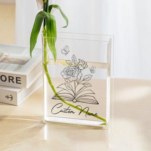 May include: A clear, rectangular vase with a floral and butterfly design, holding a red carnation. The vase has the words "Custom Name" on it. The vase is on a light-colored surface, with books in the background.