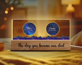 May include: A rectangular light-up plaque with a wooden base featuring the text The day you became our dad. The acrylic panel displays two circular star maps labeled Max 24/10/2009 and Emma 24/05/2005 above a dark blue mountain silhouette.