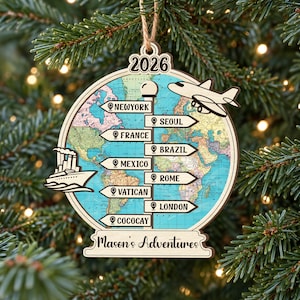 May include: A round wooden Christmas ornament featuring a world map background, a central signpost with arrows pointing to New York, Seoul, France, Brazil, Mexico, Rome, Vatican, London, and Cococay. It includes a plane, a ship, the year 2026, and the text Mason's Adventures.
