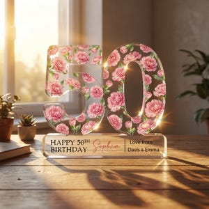 May include: A clear acrylic '50' with pink floral design, standing on a base with text that reads 'Happy 50th Birthday Sophia'. The base also includes 'Love from Davis & Emma'. The numbers are illuminated, creating a celebratory feel.