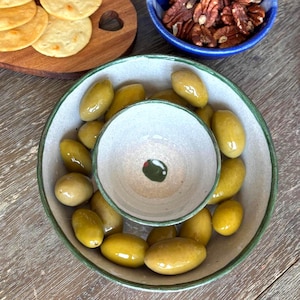 May include: A selection of appetizers, including olives, crackers, and pecans, arranged on a wooden surface. The olives are in a two-tiered ceramic bowl, and the crackers are on a wooden serving board with a heart-shaped cutout. A blue ceramic bowl and a blue ceramic jar are also visible.