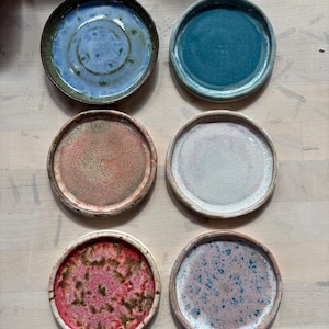 May include: Six circular ceramic coasters arranged in two columns. The top left is blue with a spiral, the top right is solid teal. The middle left is reddish-brown, the middle right is off-white. The bottom left is bright pink with dark speckles, the bottom right is light pink with blue speckles.