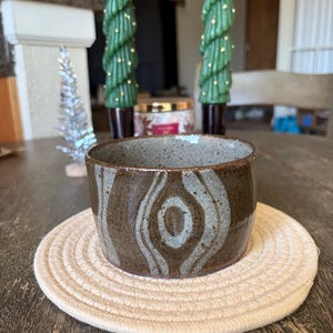 May include: A handmade ceramic bowl with a brown and gray swirl design, resting on a beige woven coaster. The bowl has a rustic, textured appearance. Decorative green tree-shaped candles are in the background.