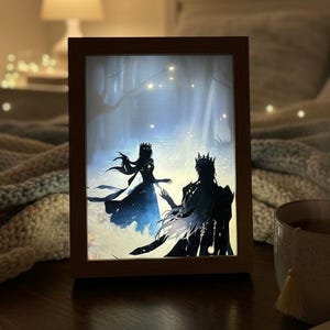 Può includere: Una scatola luminosa con cornice in legno con un'illustrazione silhouette di una coppia in un ambiente forestale. L'opera raffigura un uomo e una donna con corone. Una tazza di tè e un libro aperto sono sul tavolo.