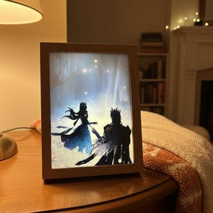 Può includere: Una stampa artistica incorniciata raffigurante due figure silhouette, forse un re e una regina, in un ambiente forestale. L'opera d'arte utilizza tonalità blu e nere, con una cornice in legno. La scena è illuminata delicatamente.