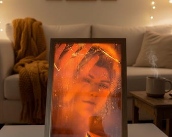 May include: A framed portrait with a warm, orange glow, featuring a person's face and hands. The frame is light-colored wood, and the image is set on a white table. The background includes a white couch, framed photos, and string lights.
