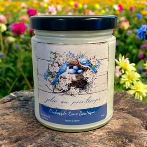 Puede incluir: Un tarro de vela de vidrio verde con una etiqueta blanca que presenta un nido con p&aacute;jaros azules y huevos. La etiqueta dice "jolie au printemps" y "Crabapple Lane Boutique". La vela est&aacute; etiquetada como "Sweet Cotton" y es de 9 oz.