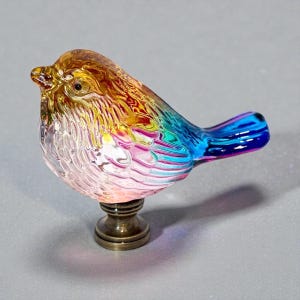 Puede incluir: Una figura de pájaro de cristal con un degradado de colores ámbar, amarillo, rosa y azul con un patrón de plumas texturizado. El pájaro está montado sobre una base metálica de color bronce, circular y escalonada, creando un remate decorativo.