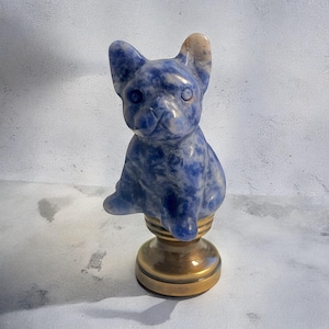 May include: A blue and white carved stone figurine of a French bulldog sitting on a gold metal base.