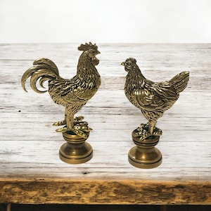 May include: Two gold metal rooster and hen figurines on a wooden surface. The rooster is standing on a round base and the hen is standing on a similar base. Both figurines have detailed features and a textured finish.