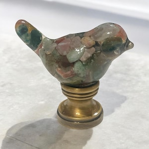 Könnte beinhalten: Ein kleiner, gläserner Vogelknauf mit Messingfuß. Der Vogel ist aus klarem Glas mit eingebetteten bunten Steinen, darunter rosa, grün und braun.