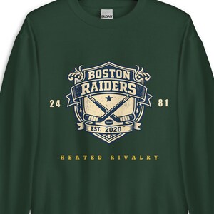 Könnte beinhalten: Ein dunkelgrünes Sweatshirt mit einem Boston Raiders-Logo. Das Logo zeigt gekreuzte Hockeyschläger, einen Stern und den Text "EST. 2020". Die Worte "BOSTON RAIDERS" sind über dem Logo angeordnet, mit "24" und "81" auf beiden Seiten. Der Satz "HEATED RIVALRY" steht darunter.