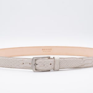 Peut inclure: Une ceinture en cuir blanc avec une boucle argentée. La ceinture est embossée d'un motif et porte l'inscription "Made in Italy" au dos.