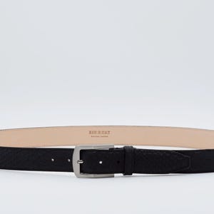 Könnte beinhalten: Ein schwarzer Ledergürtel mit einer silbernen Schnalle. Der Gürtel hat eine strukturierte Oberfläche und die Schnalle ein schlichtes Design. Der Gürtel ist in Italien hergestellt.