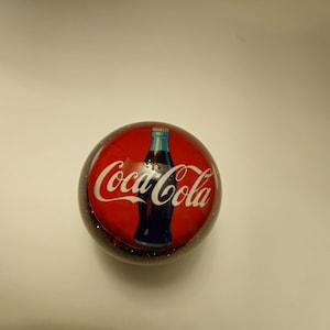 May include: A red and black round bottle stopper with a Coca-Cola logo and a bottle of Coca-Cola on it.