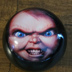 Könnte beinhalten: Ein schwarzes, rundes Dekorationsstück mit einem detaillierten Bild eines Puppengesichts. Die Puppe hat blaue Augen, rotes Haar und einen finsteren Ausdruck. Das Gesicht der Puppe steht im Mittelpunkt des Objekts.