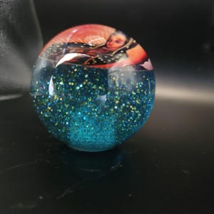 Op de afbeelding: Een heldere, bolvormige presse-papier met een blauwe glitterbasis. De bovenkant van de presse-papier heeft een afbeelding van een persoon met rode en zwarte accenten. De glitter is een mix van blauw en goud.