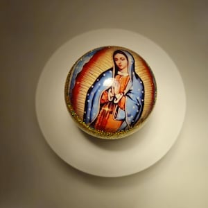 May include: A round, gold-coloured ornament featuring a picture of the Virgin Mary in a blue robe. The ornament has a white background with gold glitter.