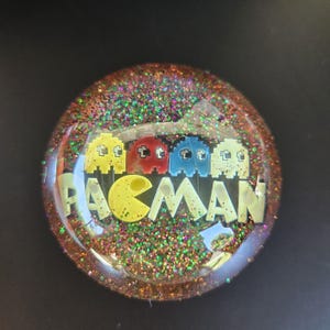 Può includere: Decorazione rotonda a tema Pac-Man, piena di glitter. Il design presenta la parola "PACMAN" in giallo, insieme a personaggi pixelati di Pac-Man in giallo, rosso e blu. Lo sfondo è un mix di glitter.