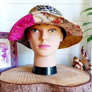 May include: A wide-brimmed sun hat made from patchwork fabric in shades of pink, yellow, brown, and blue. The hat has a floral pattern and is worn on a mannequin head.
