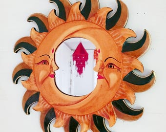 May include: A wooden sun and moon mirror with a green and gold painted design. The mirror is round and has a face on each side. The sun has a yellow face with a red mouth and the moon has a white face with a red mouth.