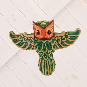 以下が含まれることがあります： 木製のフクロウの置物。羽を広げたデザインで、緑、オレンジ、タンの色合いで塗装されています。羽は大きく広げられ、フクロウの目は暗いです。この置物は装飾品です。