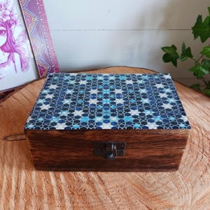 Puede incluir: Una caja rectangular de madera con un acabado teñido en marrón oscuro y un cierre de pestillo metálico. La tapa está decorada con un vibrante patrón de mosaico geométrico azul y blanco que consiste en formas de estrellas y polígonos.