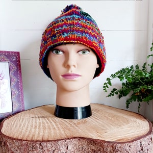 May include: A colorful knitted beanie with a pom pom on top. The beanie is red, orange, yellow, green, blue, and purple. The beanie is on a wooden stand.