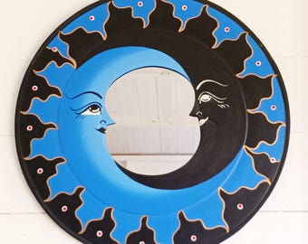 May include: Round wall mirror featuring a celestial design with a blue crescent moon and a black sun, each with a face. The mirror is framed with a black border, adorned with gold accents and red dots. A unique decorative piece.