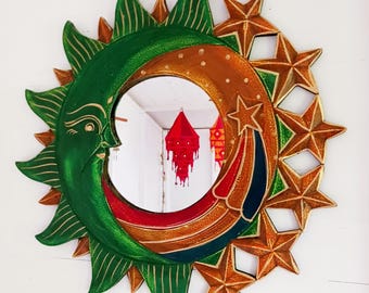 May include: A round wooden mirror with a green sun and a gold moon design. The mirror is surrounded by stars and has a rainbow-colored stripe.