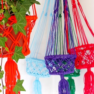 May include: A selection of vibrant macrame plant hangers in red, blue, purple, and green. Each hanger has a woven basket design with a tassel at the base, ideal for showcasing potted plants. The hangers are displayed against a white backdrop.