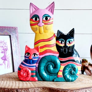 May include: A hand-painted wooden sculpture featuring three cats of varying sizes. The tallest cat is yellow with pink and red stripes, the medium cat is black with colourful stripes, and the smallest cat is red with blue and pink stripes.