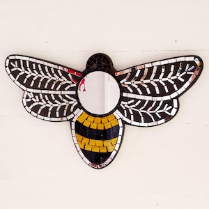 May include: A bee-shaped mosaic mirror with a circular central mirror. The wings are black with white and silver details, the body is black and yellow, and the head is black. It is mounted on a white surface.