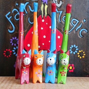 Peut inclure: Quatre figurines de chat en bois peintes à la main, hautes et élancées, de couleurs rouge, orange, bleue et verte. Chaque chat présente un long cou, des motifs floraux peints sur le corps, un visage blanc avec des moustaches et une petite clochette.