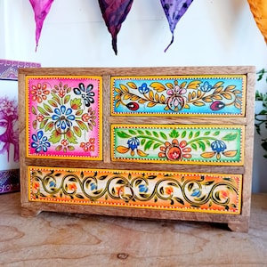 Op de afbeelding: Een kleine houten ladekast met vier lades. Elke lade is handbeschilderd met kleurrijke bloemmotieven in roze, blauw, oranje en groen. De kast heeft een natuurlijke houten afwerking en een decoratief bloemmotief op de voorkant.