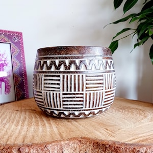 Op de afbeelding: Een donkerbruine houten plantenbak met een gesneden geometrisch ontwerp. De plantenbak heeft een cilindrische vorm met een brede opening en is versierd met witte, gesneden patronen. De plantenbak staat op een houten oppervlak.