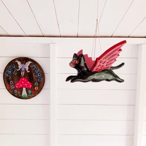 May include: A hanging black cat ornament with red wings and orange eyes, suspended by thin strings. Beside it, a circular wooden sign features a painted red mushroom, a small fairy figure, and the text Fairy magic in blue lettering.