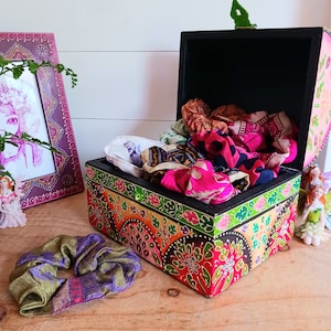 May include: A colourful wooden box with a black lid and intricate floral designs. The box is open and filled with colourful fabric scraps.