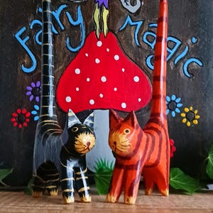 Puede incluir: Figuras de gato de madera talladas a mano en negro y naranja con detalles dorados. Los gatos están frente a un letrero de madera con las palabras "Fairy Magic" y una seta roja con lunares blancos. También hay detalles florales decorativos.