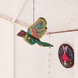 May include: A green, wooden, flying creature with red eyes and multi-coloured wings is suspended from a black string. The wings have pink, yellow, and blue accents. A round sign with the words "fairy magic" is visible in the background.