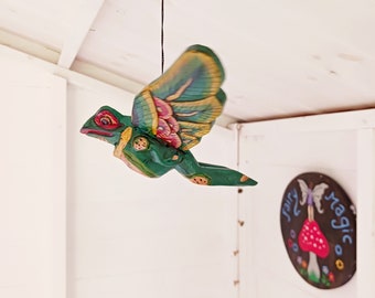 May include: A green, wooden, flying creature with red eyes and multi-colored wings is suspended from a black string. The wings have pink, yellow, and blue accents. A round sign with the words "fairy magic" is visible in the background.