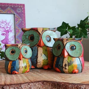 May include: Three wooden owls painted in a colorful, striped pattern. The owls have large, round eyes with green pupils and orange beaks. The owls are arranged in a row, with the largest owl in the back and the smallest owl in the front.