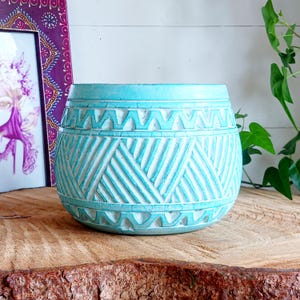 Può includere: Un vaso di ceramica turchese con un design geometrico intagliato. Il vaso ha una forma cilindrica con un'ampia apertura ed è decorato con accenti bianchi. Il vaso è appoggiato su una superficie di legno.