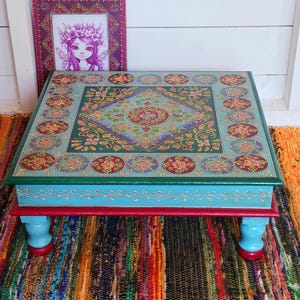 May include: A square wooden low table featuring a hand-painted surface with a central diamond motif, floral patterns, and circular border designs in shades of teal, red, green, and gold. The table has light blue sides and four turned legs.